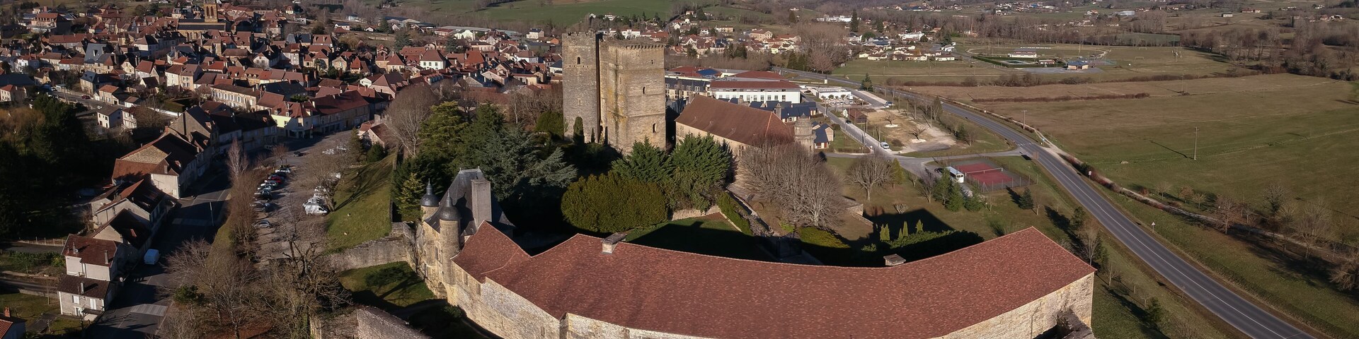 Vista aérea de un pueblo histórico con un castillo de piedra imponente, rodeado de edificios residenciales y un cielo azul despejado.