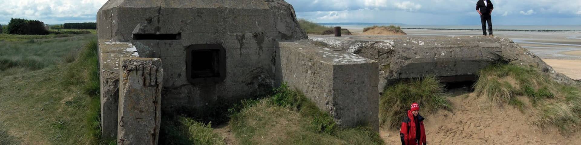A concrete bunker with a small entrance, a sandy beach, and a person standing on a raised platform.