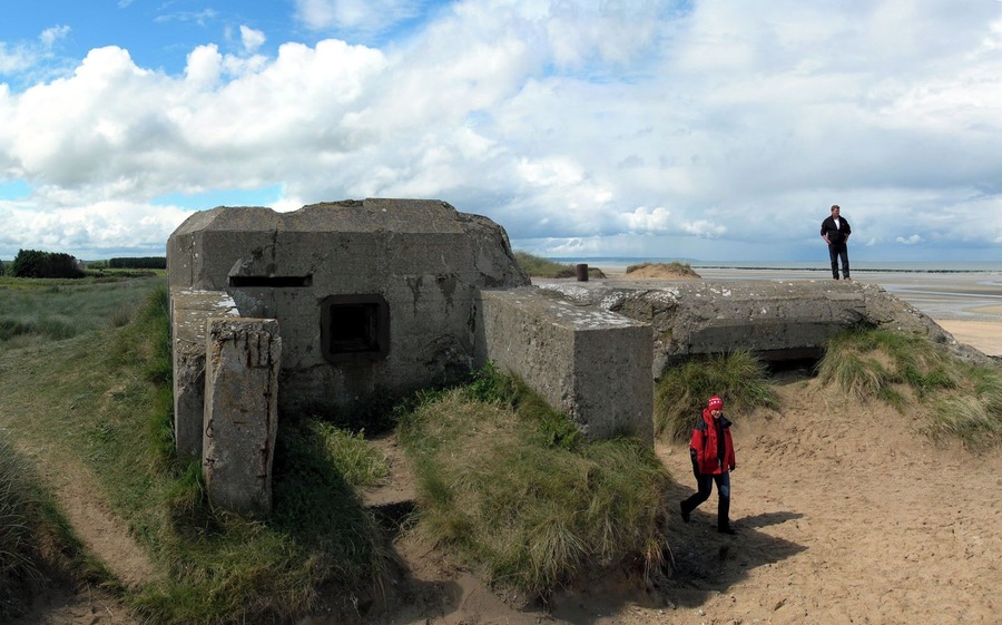 A concrete bunker with a small entrance, a sandy beach, and a person standing on a raised platform.