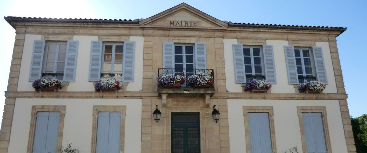 A two-story building with the sign "MAIRIE" above the entrance. It has blue shutters, flower boxes on the windows, and a small balcony with a railing.