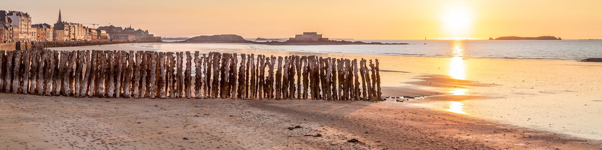 A beach with a wooden fence, a city skyline, and a sunset over the water.