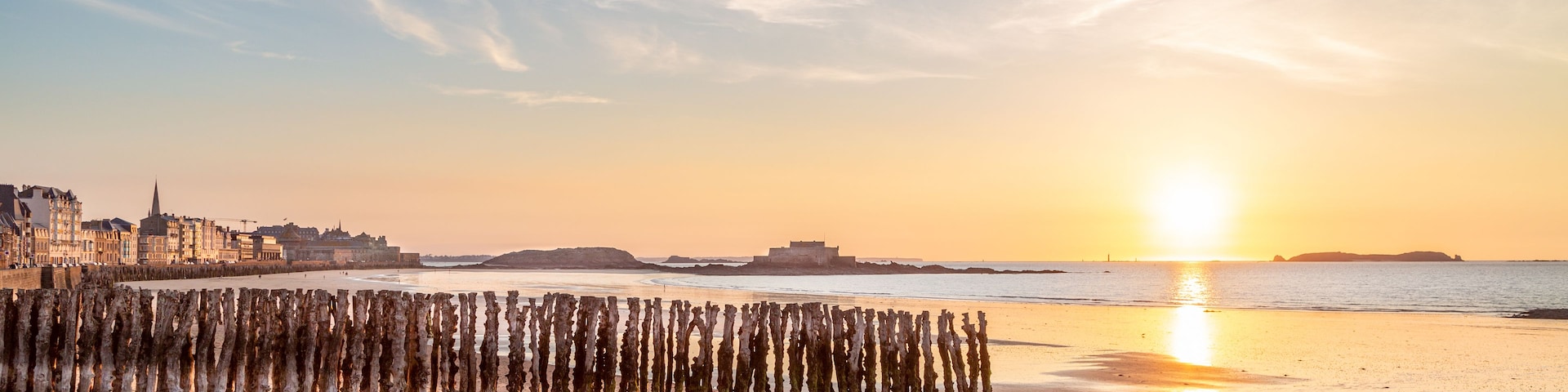 Una playa con una cerca de madera, el perfil urbano de una ciudad y una puesta de sol sobre el agua.