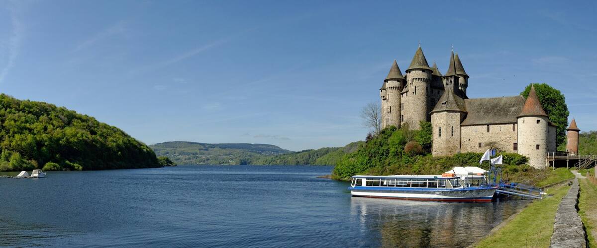A castle by a calm river with boats docked, surrounded by lush greenery.