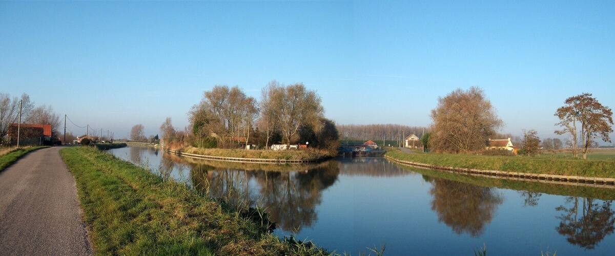 A calm river with a narrow path alongside, surrounded by trees and small buildings.