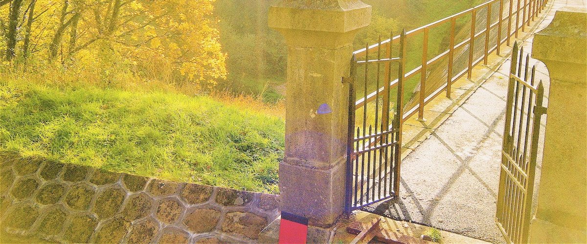 A stone bridge with a metal railing, surrounded by trees with yellow leaves, and a body of water in the background.