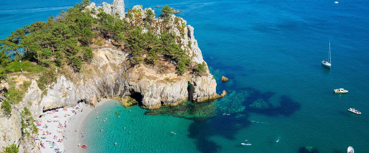 A coastal landscape with a rocky cliff, clear turquoise waters, and a beach with people sunbathing.