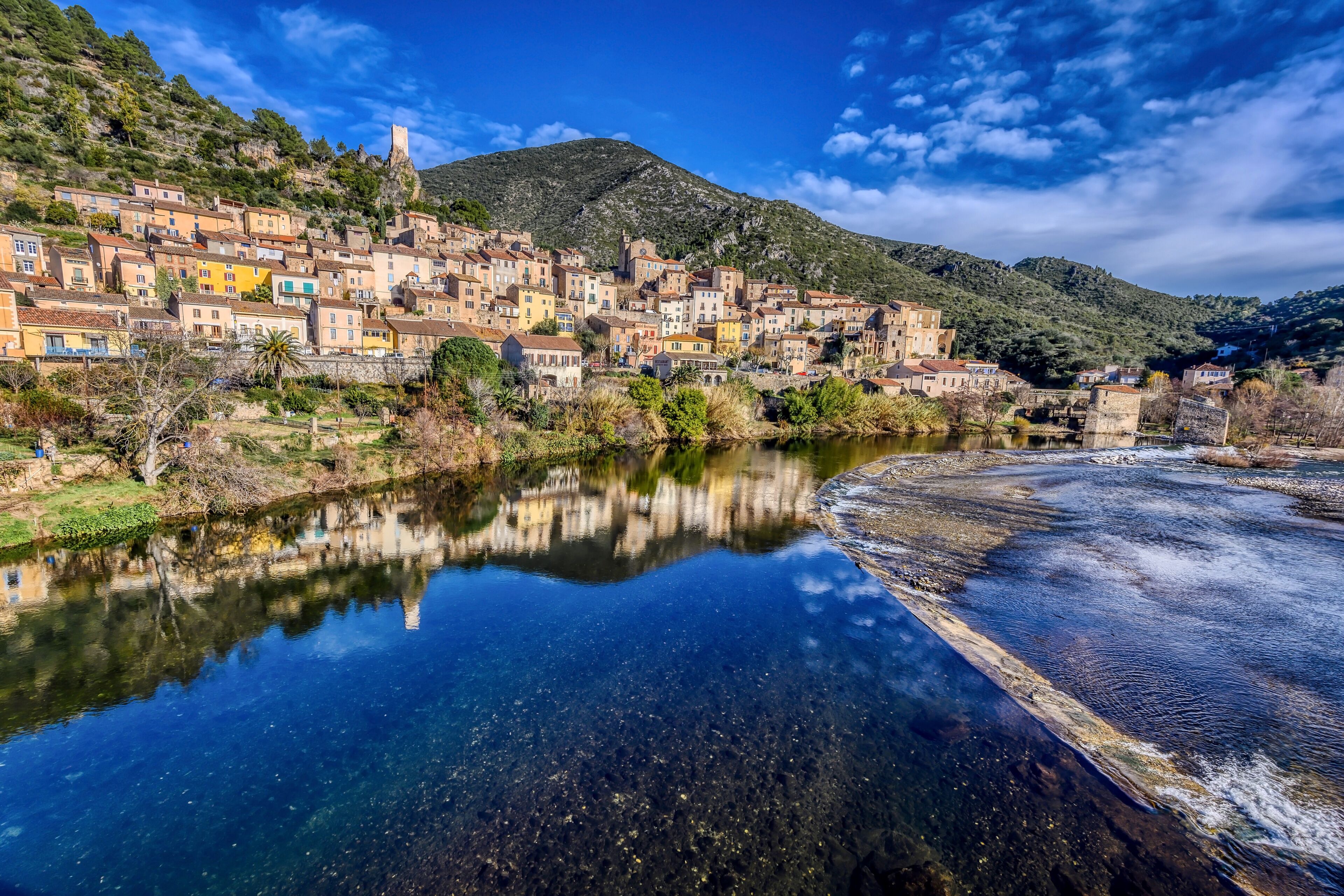 A coastal town with colorful buildings, a river reflecting the structures, and a hill in the background.