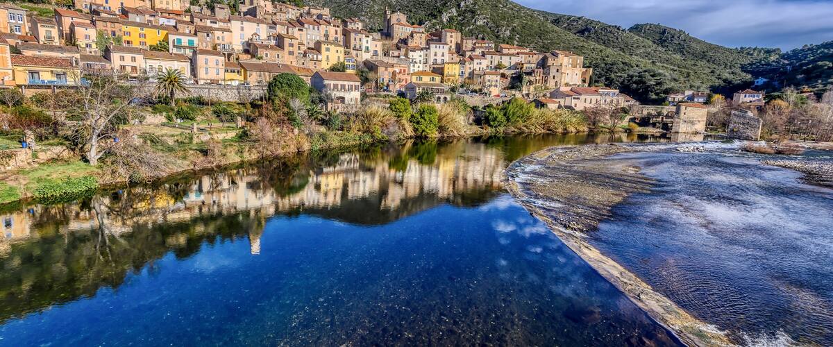 A coastal town with colorful buildings, a river reflecting the structures, and a hill in the background.