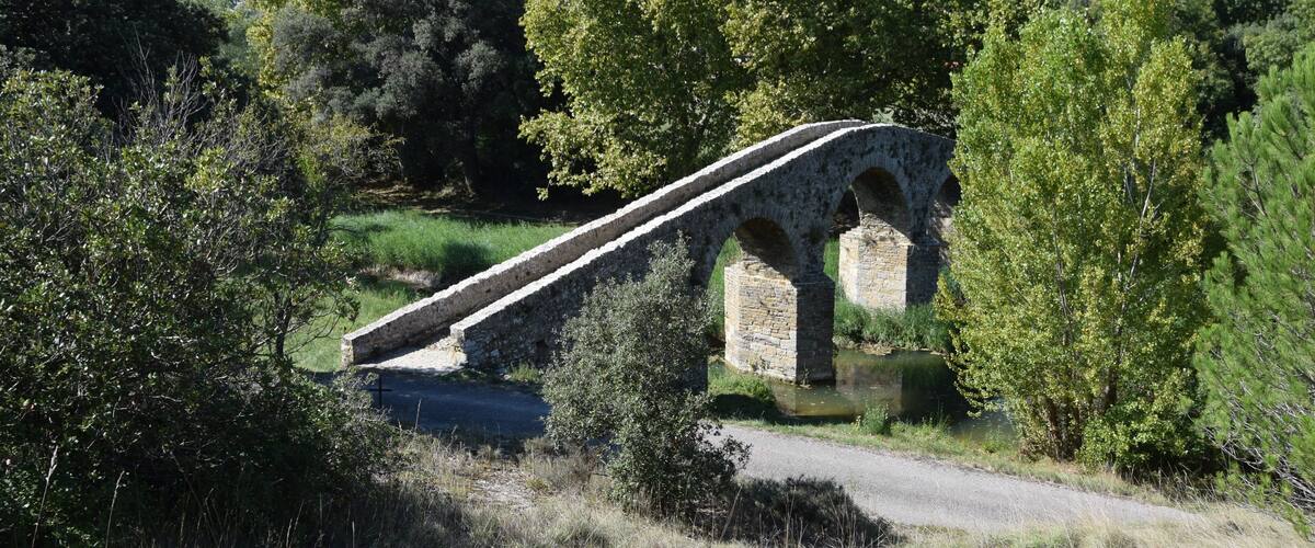 An old stone bridge with arches over a road, surrounded by greenery and trees.