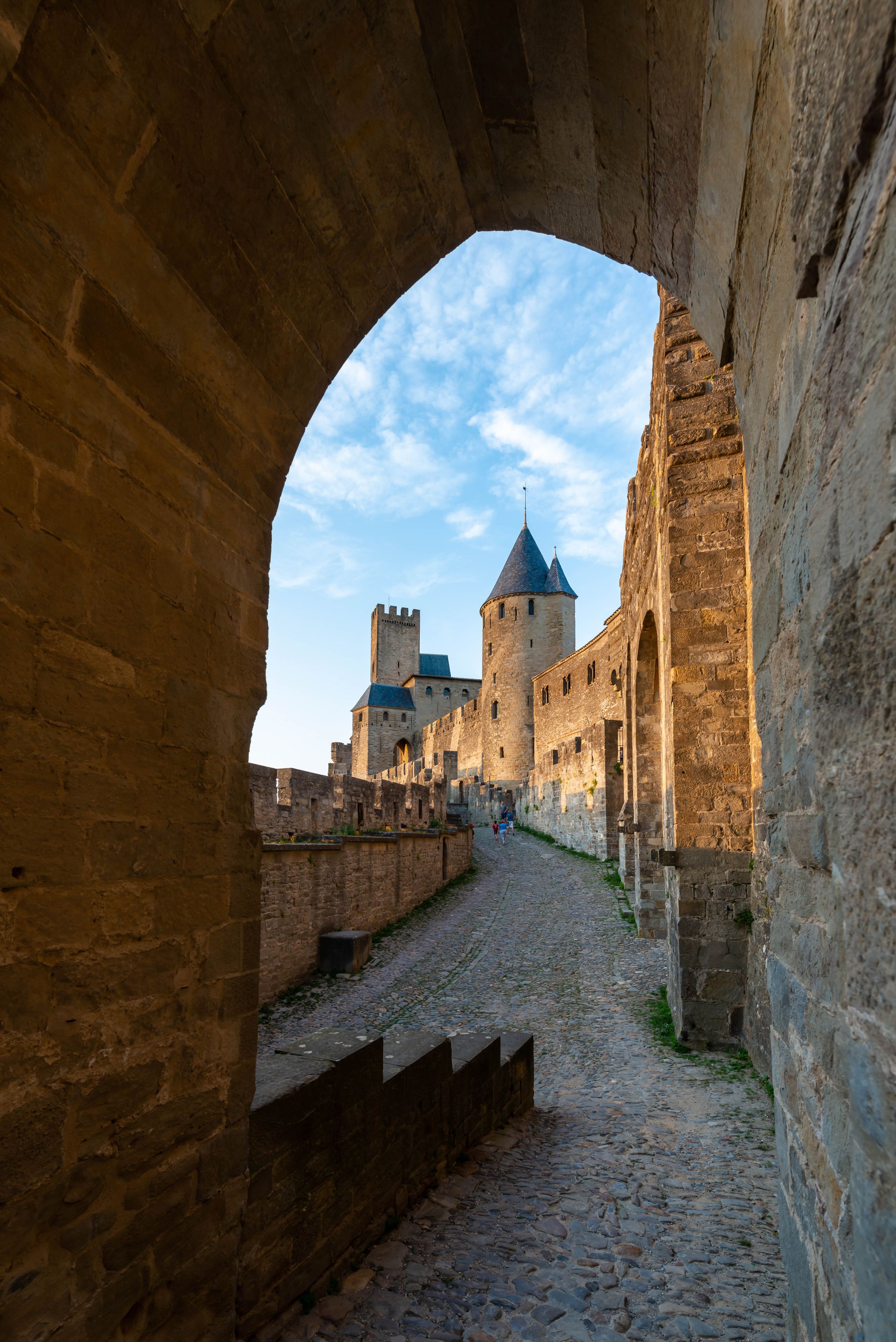 A stone archway leading to a castle with a cobblestone path and a clear sky.