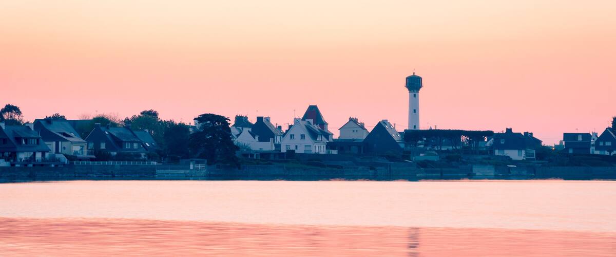 A coastal town at sunset with houses and a lighthouse.