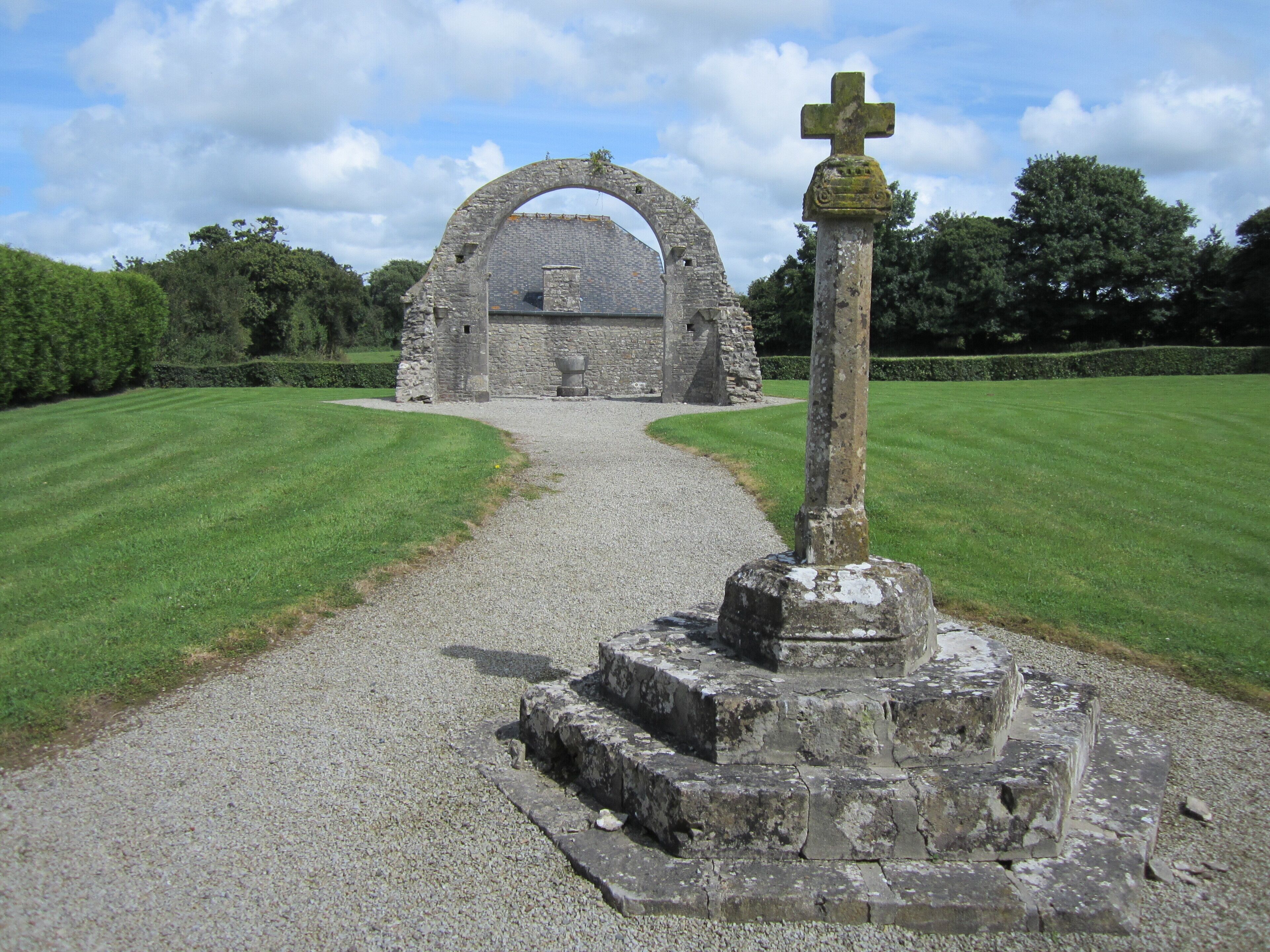 A stone archway leading to a ruined structure, surrounded by well-maintained grass and a cross at the entrance.