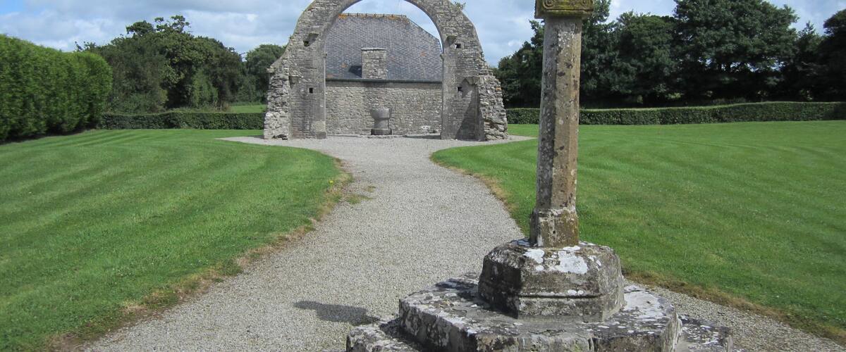 A stone archway leading to a ruined structure, surrounded by well-maintained grass and a cross at the entrance.