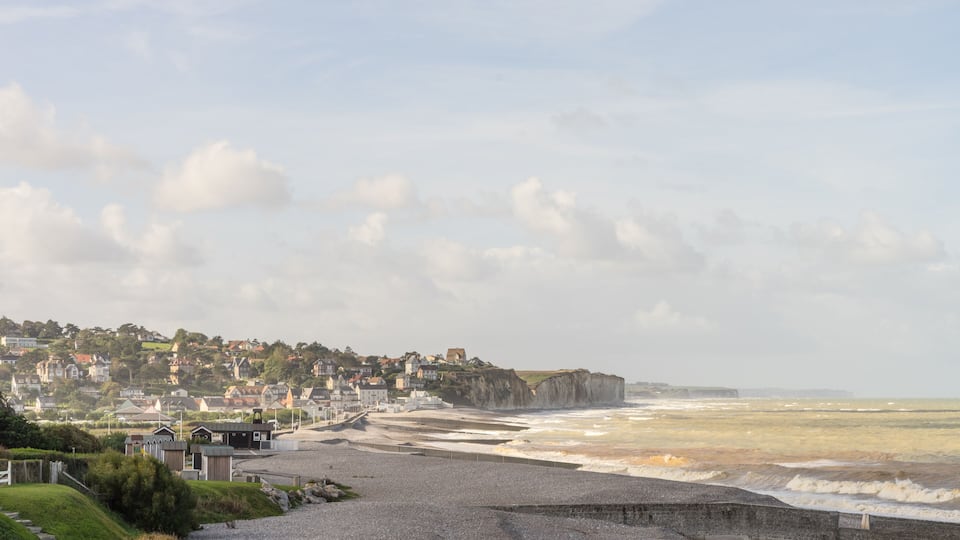 A coastal town with houses along the cliff, a pebbly beach, and waves crashing onto the shore.