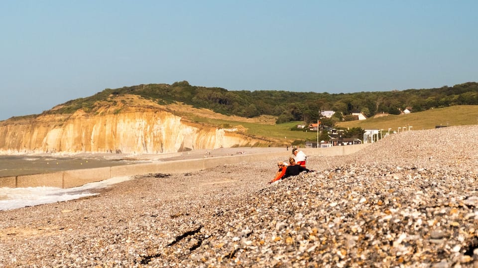 A pebbly beach with a cliff in the background and a few houses.