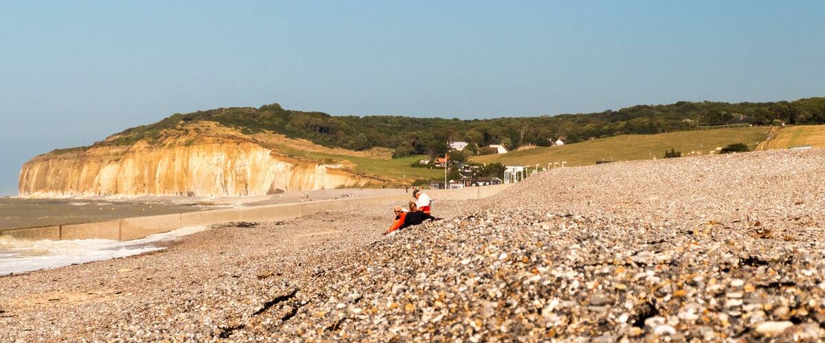 A pebbly beach with a cliff in the background and a few houses.