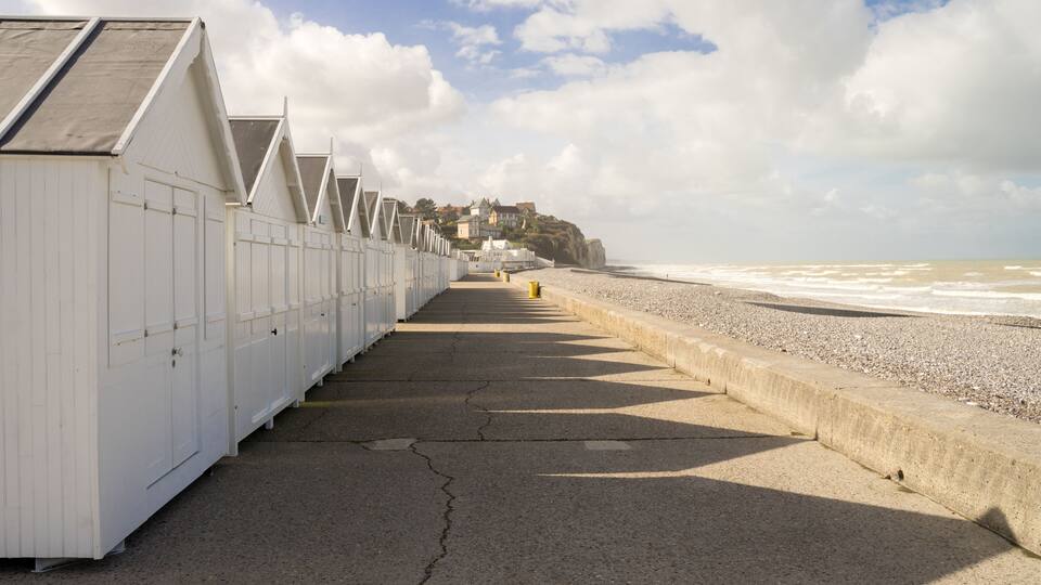 A seaside promenade with white buildings, a concrete walkway, and a beach in the background.
