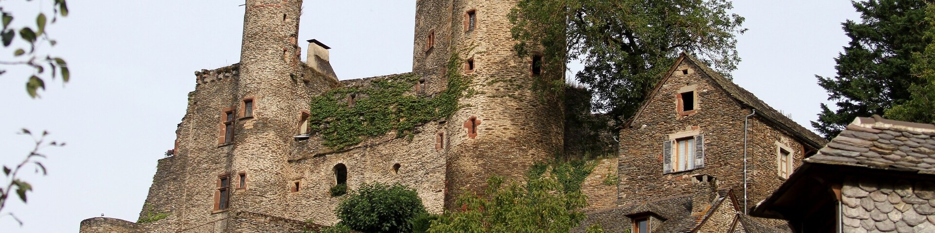 A historic stone castle with multiple towers and battlements, surrounded by lush greenery and traditional stone buildings.
