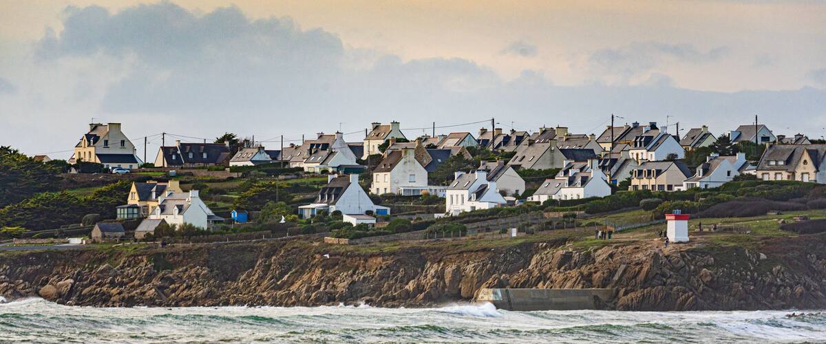 A coastal village with houses on a hillside, waves crashing onto the shore, and a lighthouse in the distance.