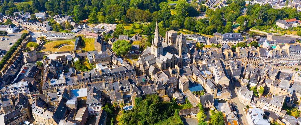 An aerial view of a historic town with medieval architecture, including a prominent church with a tall spire, surrounded by narrow streets and lush greenery.