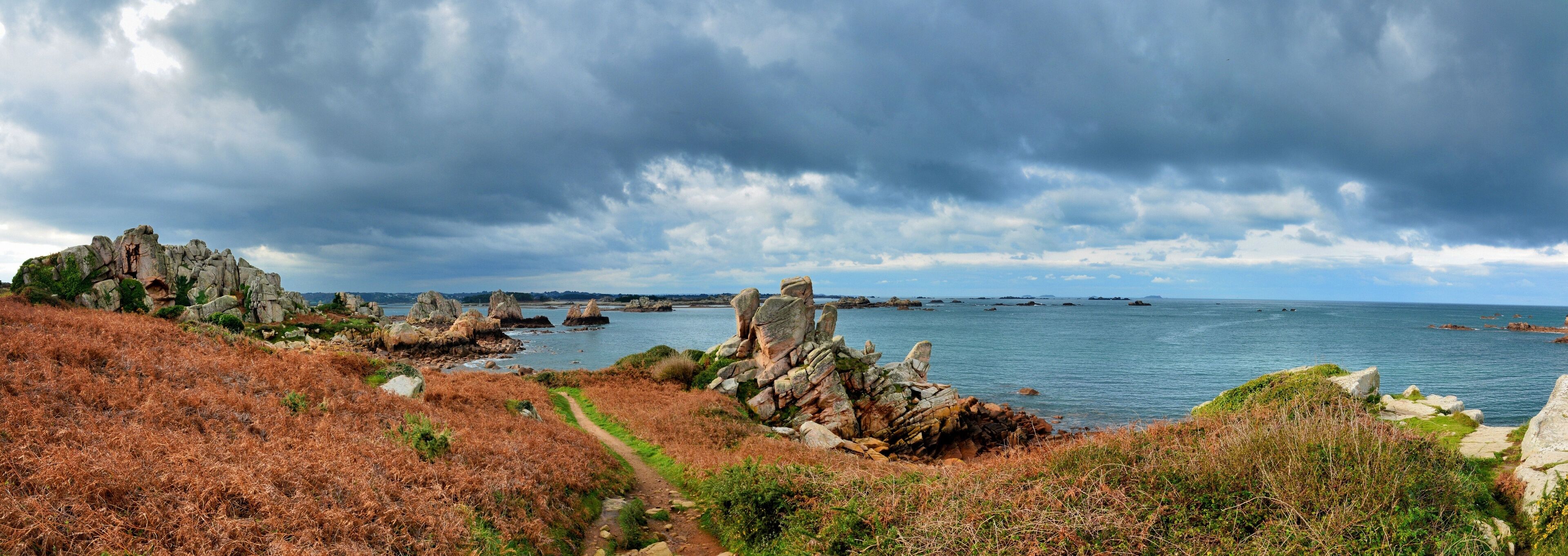 A coastal landscape with rocky outcrops, a path, and a view of the sea under a cloudy sky.