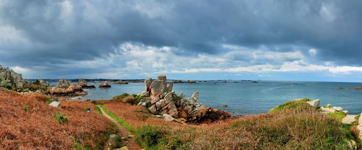 A coastal landscape with rocky outcrops, a path, and a view of the sea under a cloudy sky.
