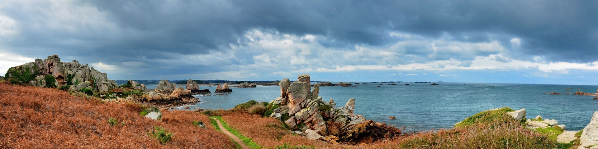 A coastal landscape with rocky outcrops, a path, and a view of the sea under a cloudy sky.