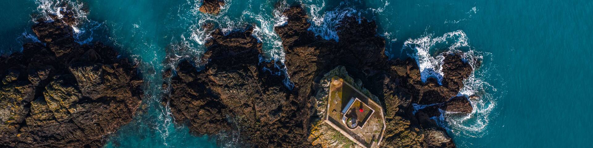 Aerial view of Phare de L'Ost-Pic lighthouse on a rocky islet with crashing waves and turquoise water in Plouezec, France.
