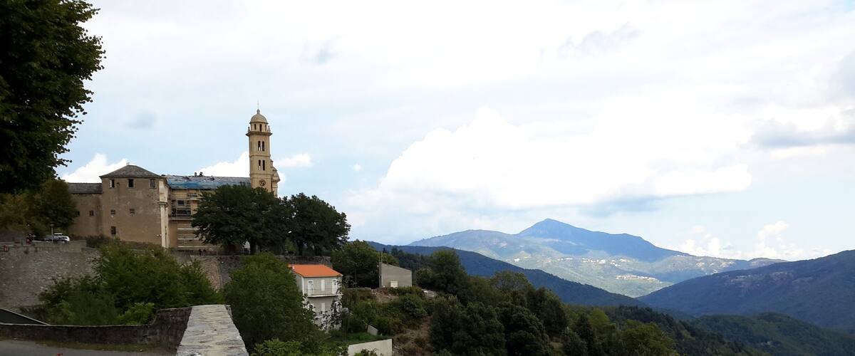 A stone wall with a dog lying on it, overlooking a mountainous landscape with a church and a tower in the background.