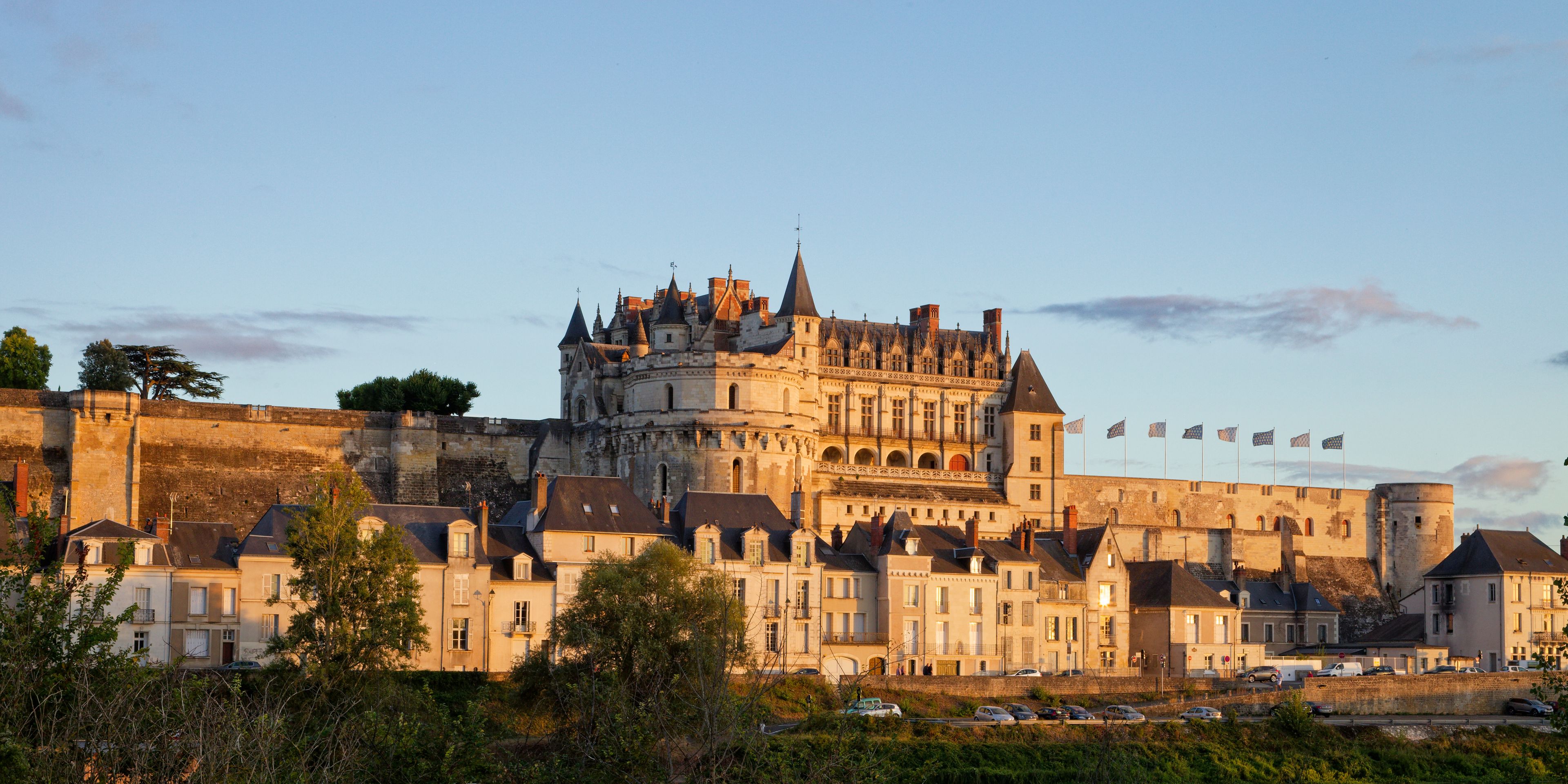 Un château historique doté de nombreuses tours et de plusieurs drapeaux, entouré d’un mur d’enceinte et bordé de bâtiments résidentiels.