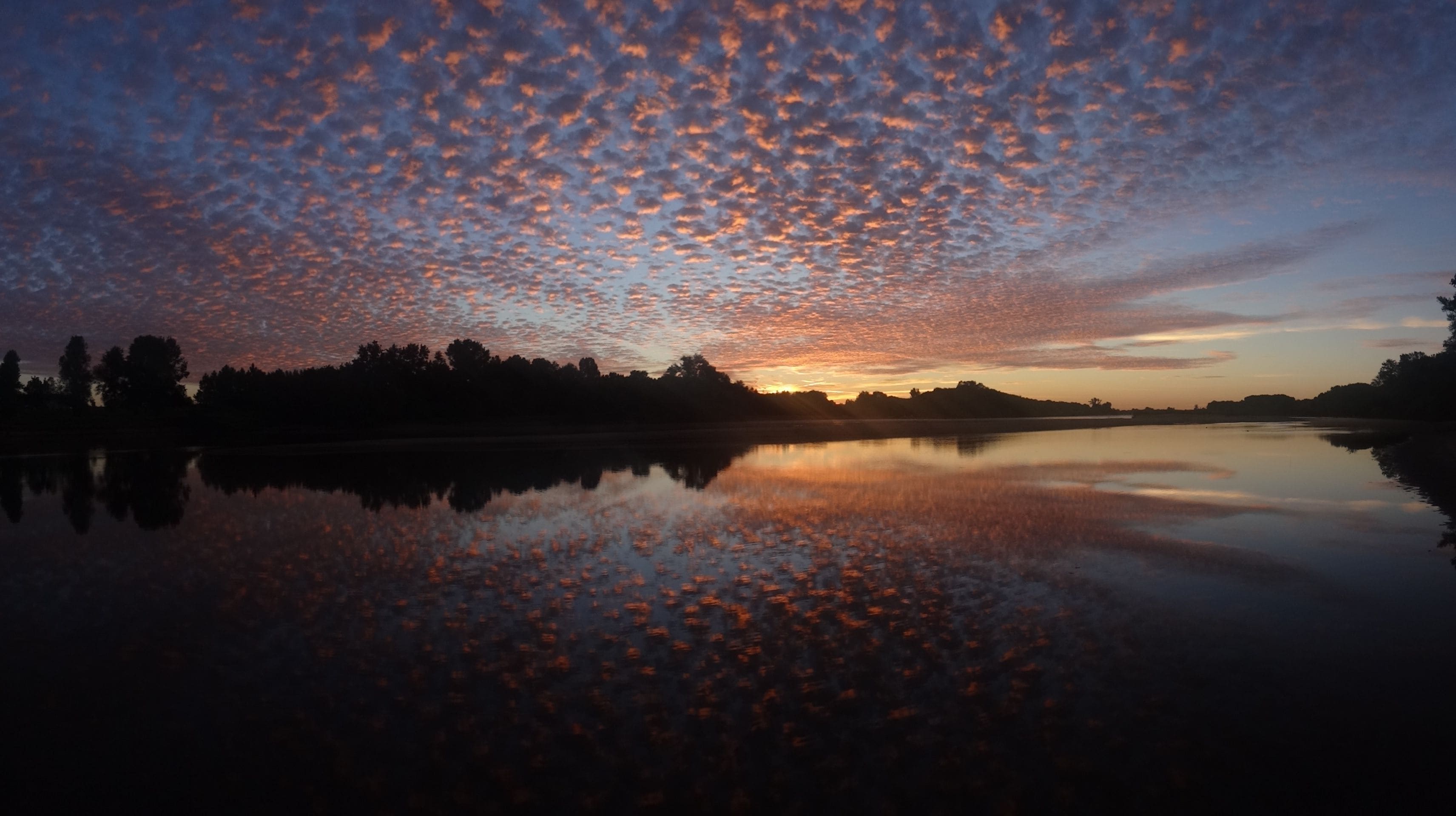 Un atardecer sobre un lago tranquilo, con la silueta de árboles y una colina distante.