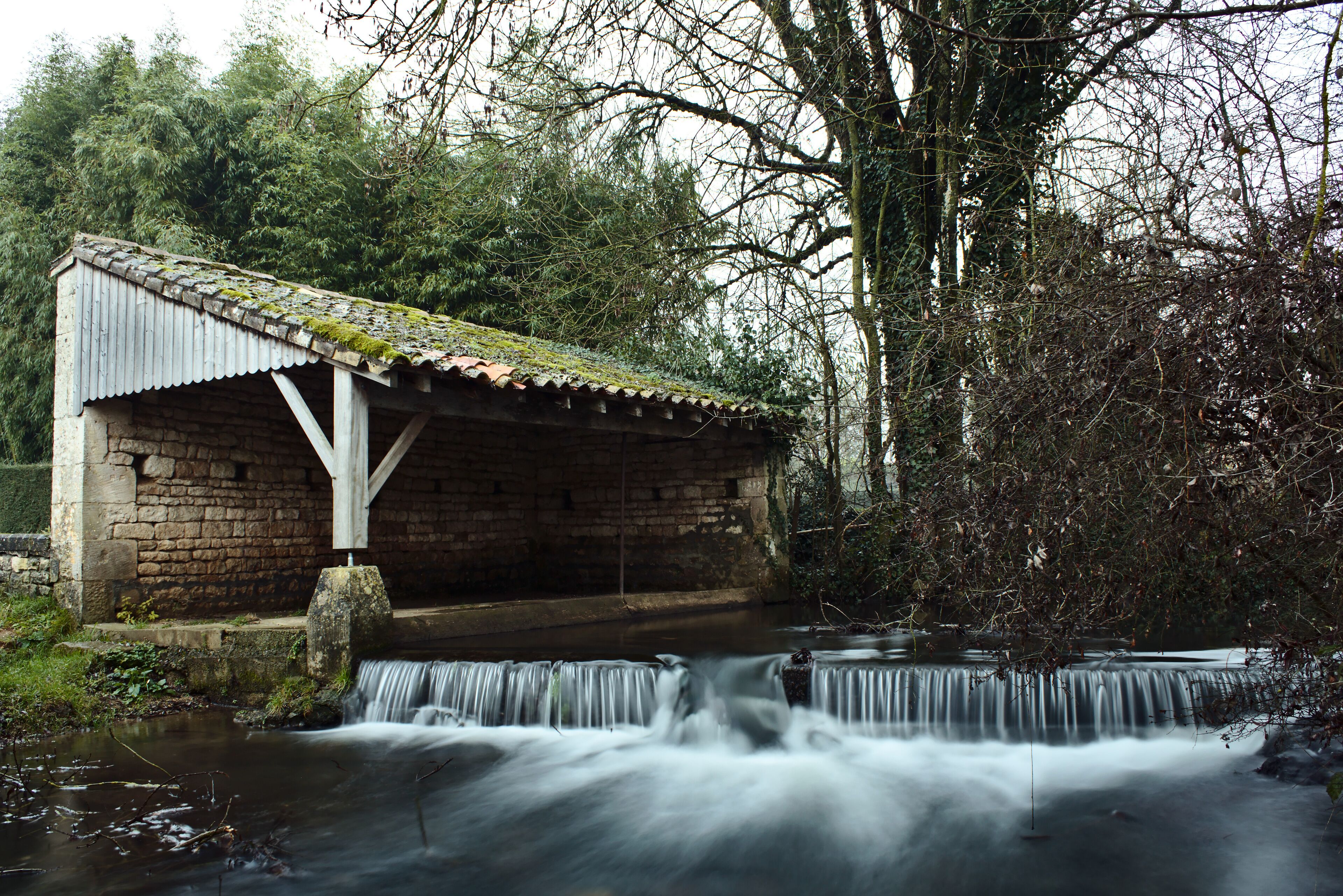 A stone bridge over a flowing stream with a small wooden shelter on the left.