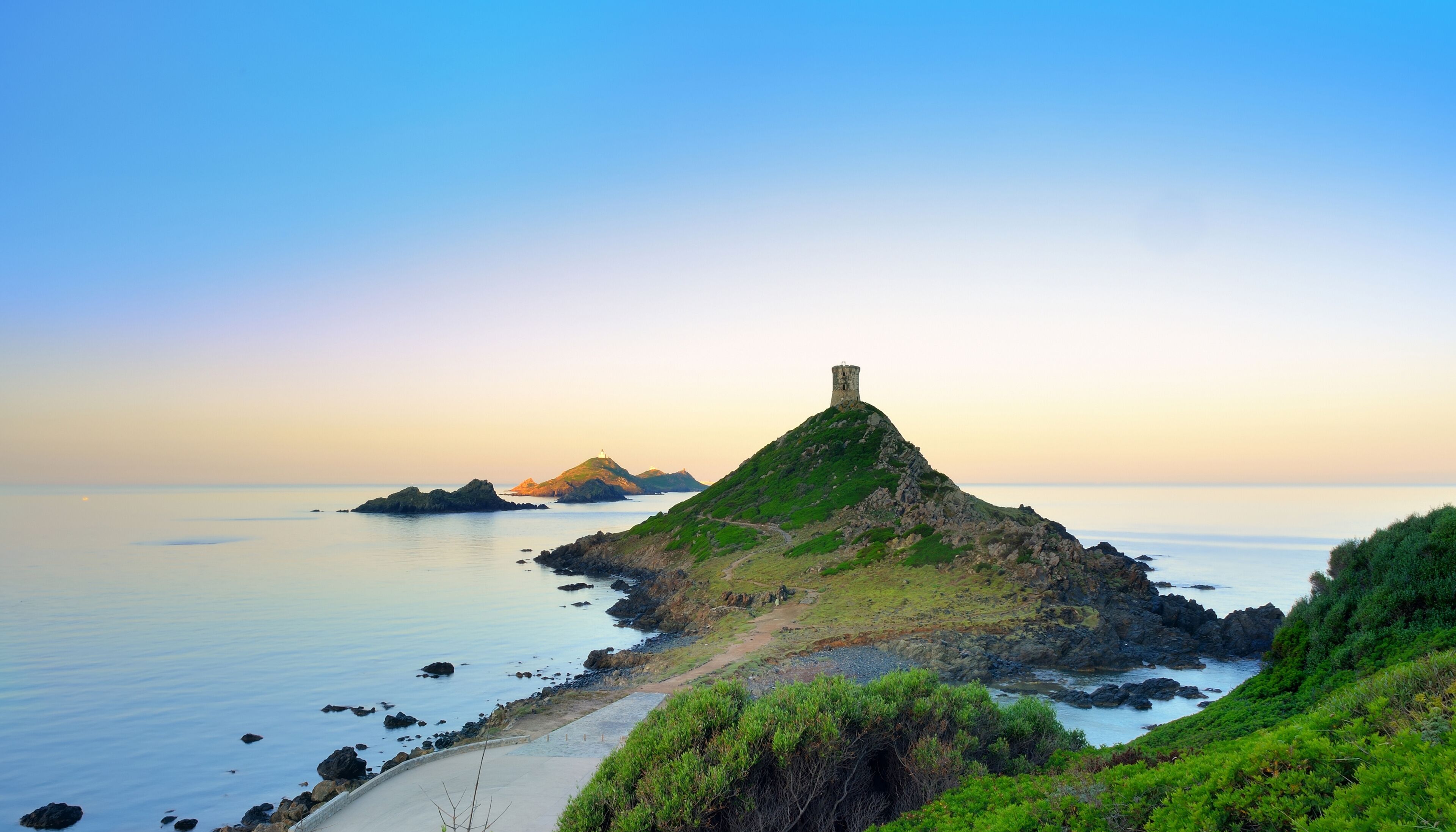 A coastal landscape with a prominent hill featuring a tower, a sandy beach, and calm waters.