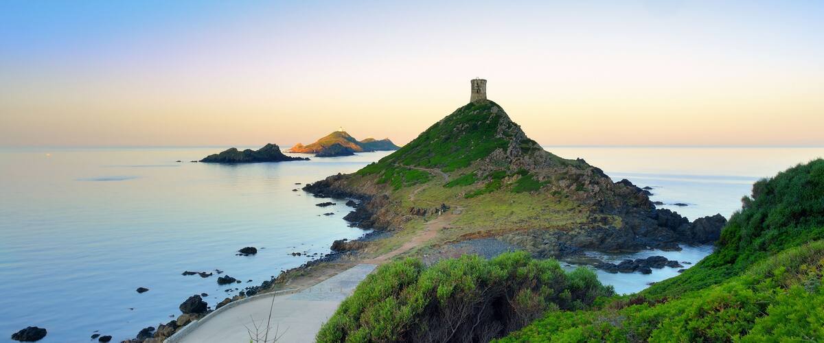 A coastal landscape with a prominent hill featuring a tower, a sandy beach, and calm waters.