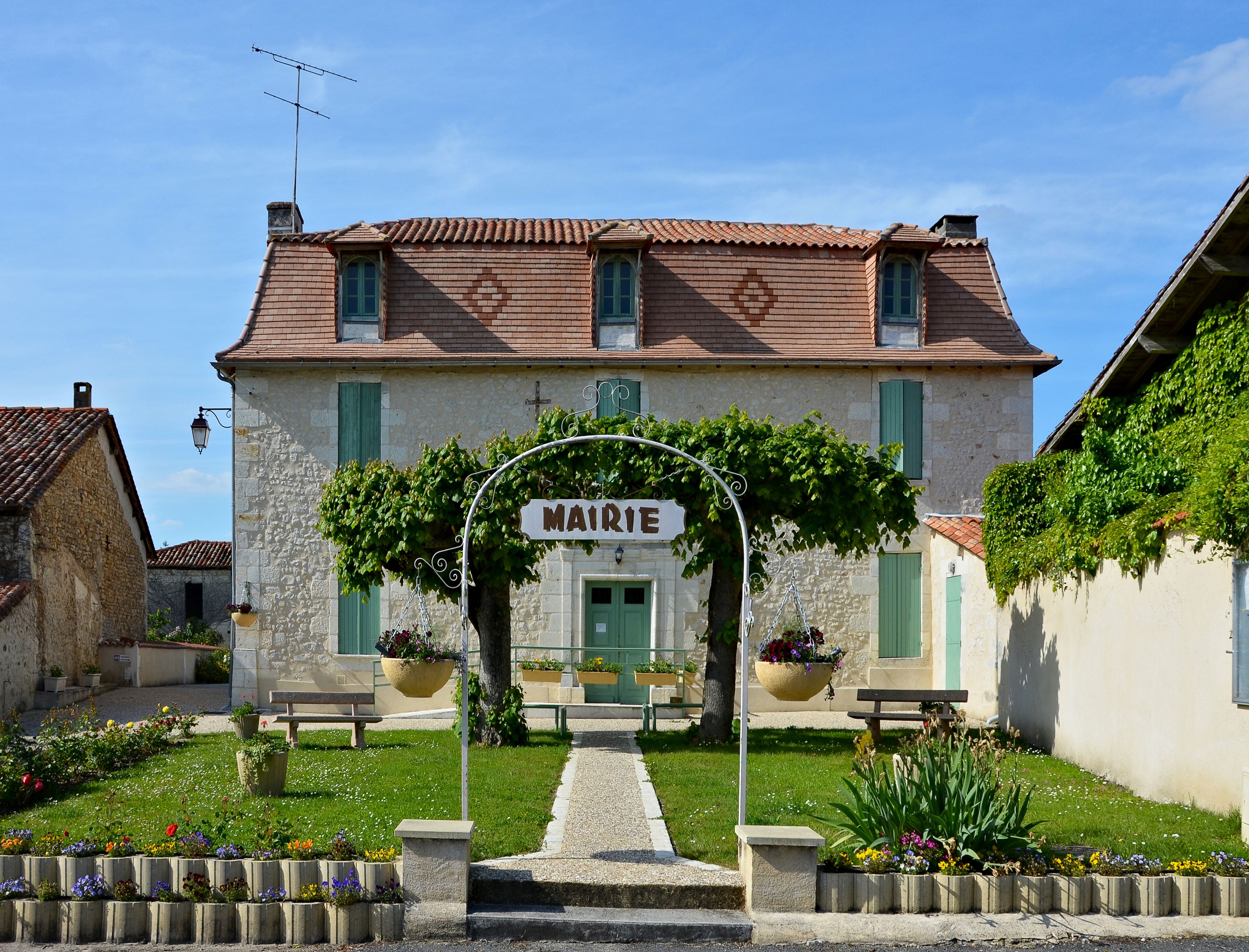 A traditional stone building with a tiled roof, a sign reading "MAIRIE," and a garden with benches and flower beds.