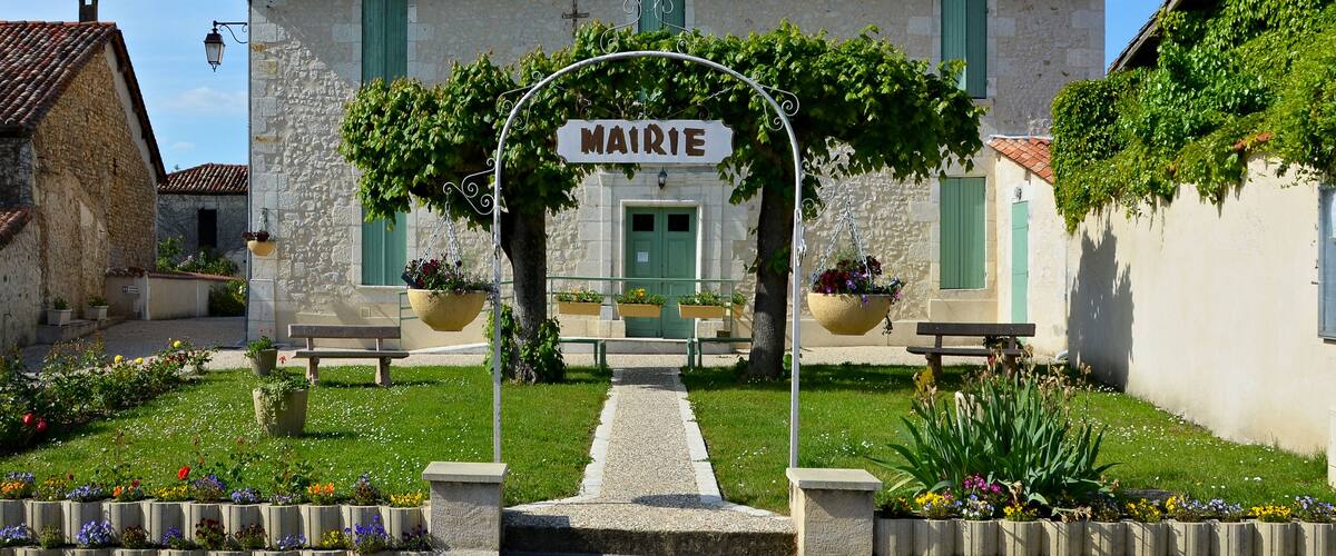 A traditional stone building with a tiled roof, a sign reading "MAIRIE," and a garden with benches and flower beds.