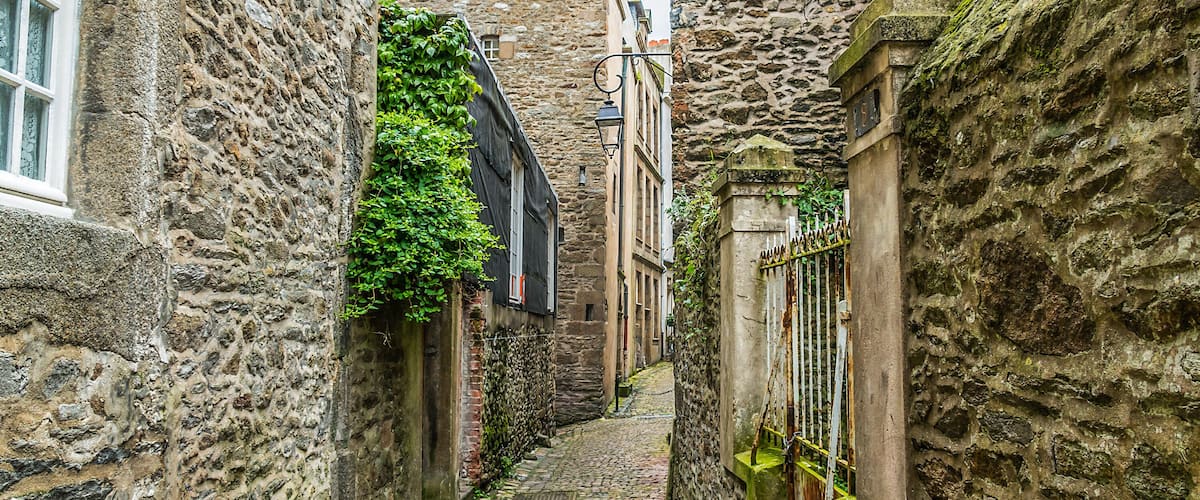 A narrow cobblestone alley flanked by stone buildings with ivy and a metal gate.