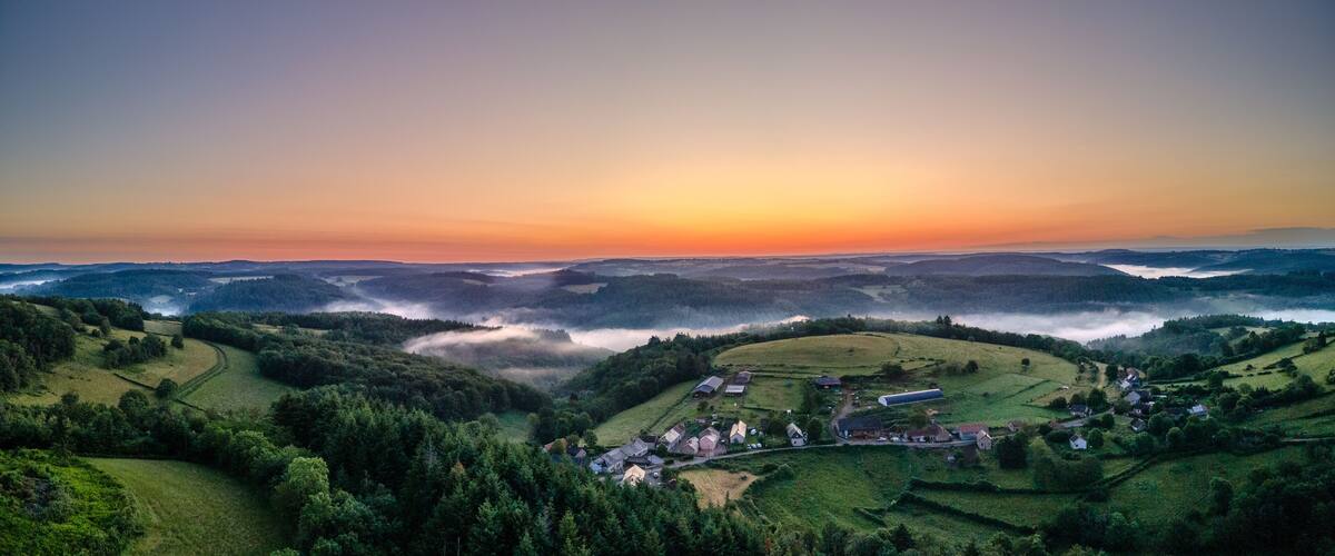 A rural landscape with rolling hills, a village, and a sunrise or sunset over the horizon.