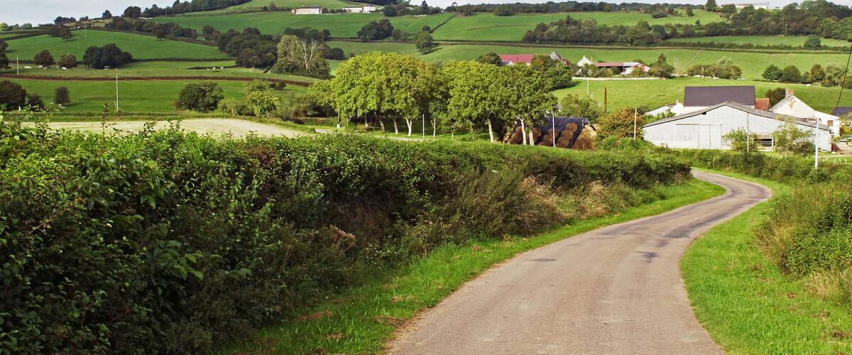 A rural road winding through green fields, with rolling hills and scattered buildings in the background.