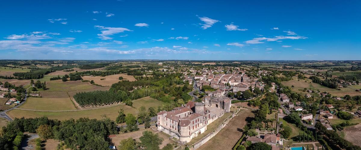 An aerial view of a historic town with a prominent castle, surrounded by green fields and a clear blue sky.