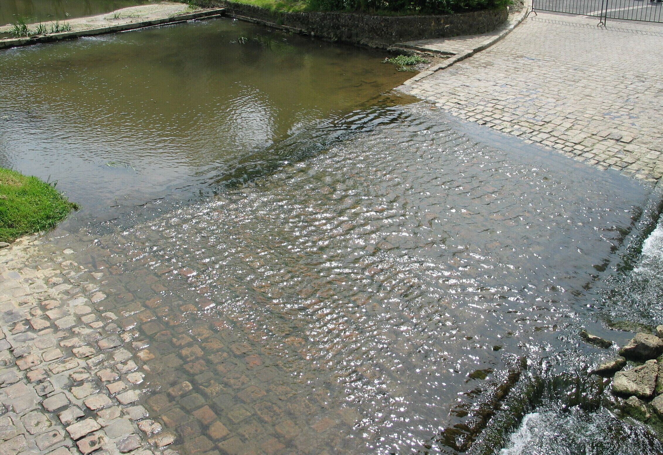 A cobblestone path alongside a calm river with a stone wall and a metal fence in the background.