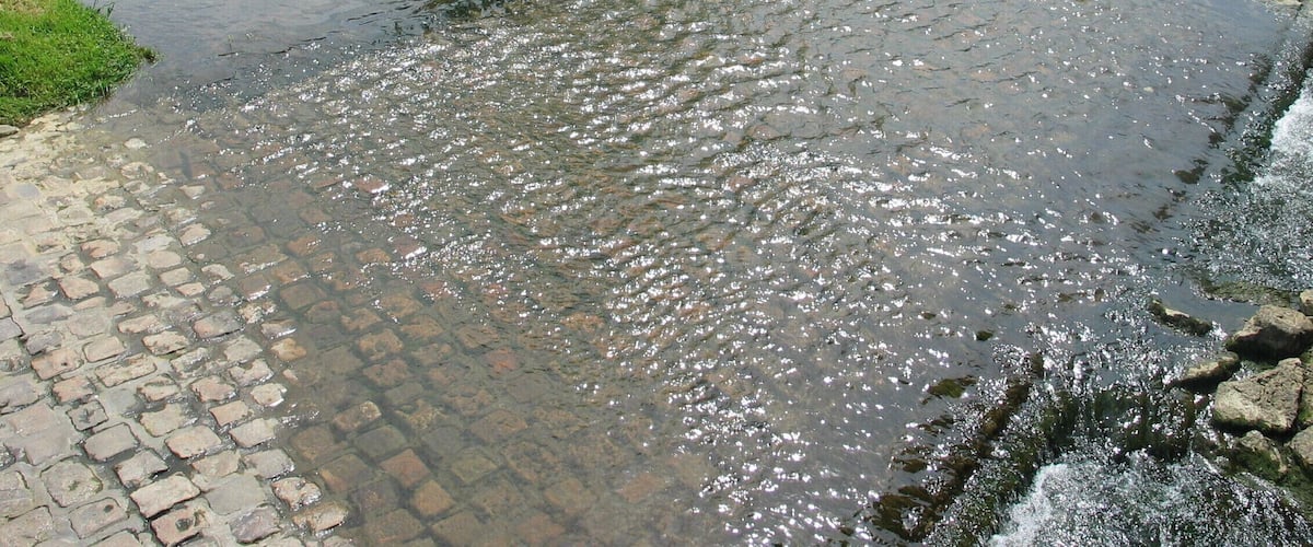 A cobblestone path alongside a calm river with a stone wall and a metal fence in the background.