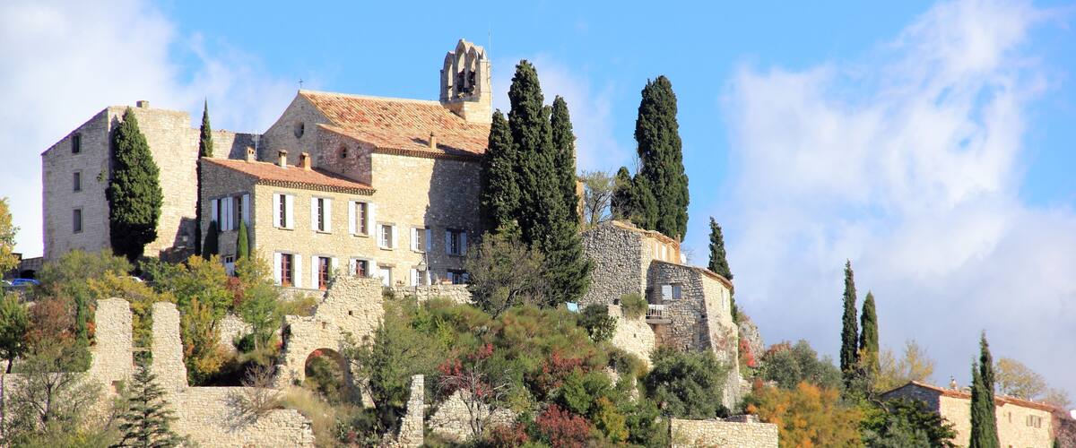 A historic stone building with a tower, surrounded by trees and ruins on a hillside.