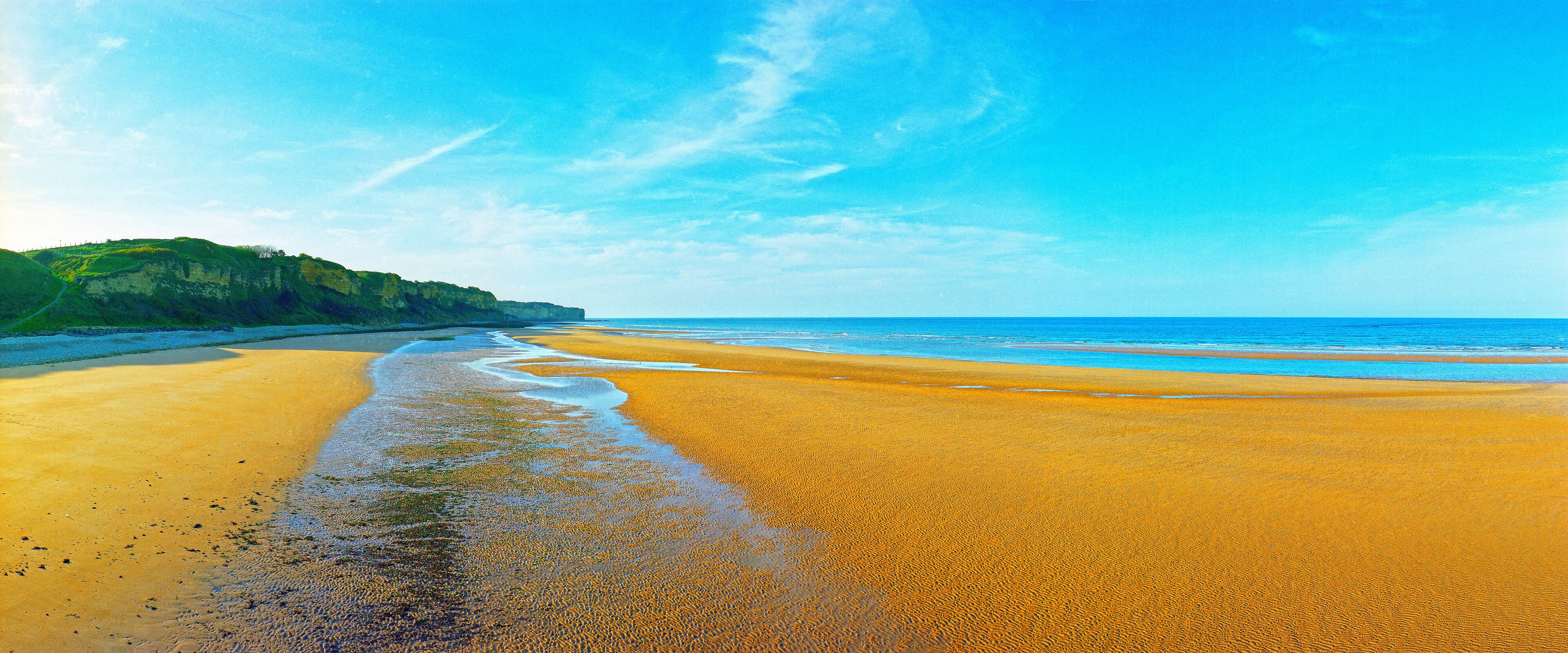 A wide sandy beach with gentle waves, a clear blue sky, and a green cliff in the background.