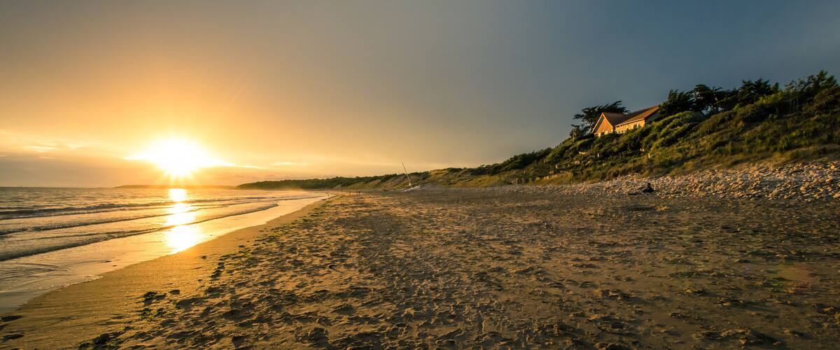 A beach with a sunset, waves, and a house on a hill.