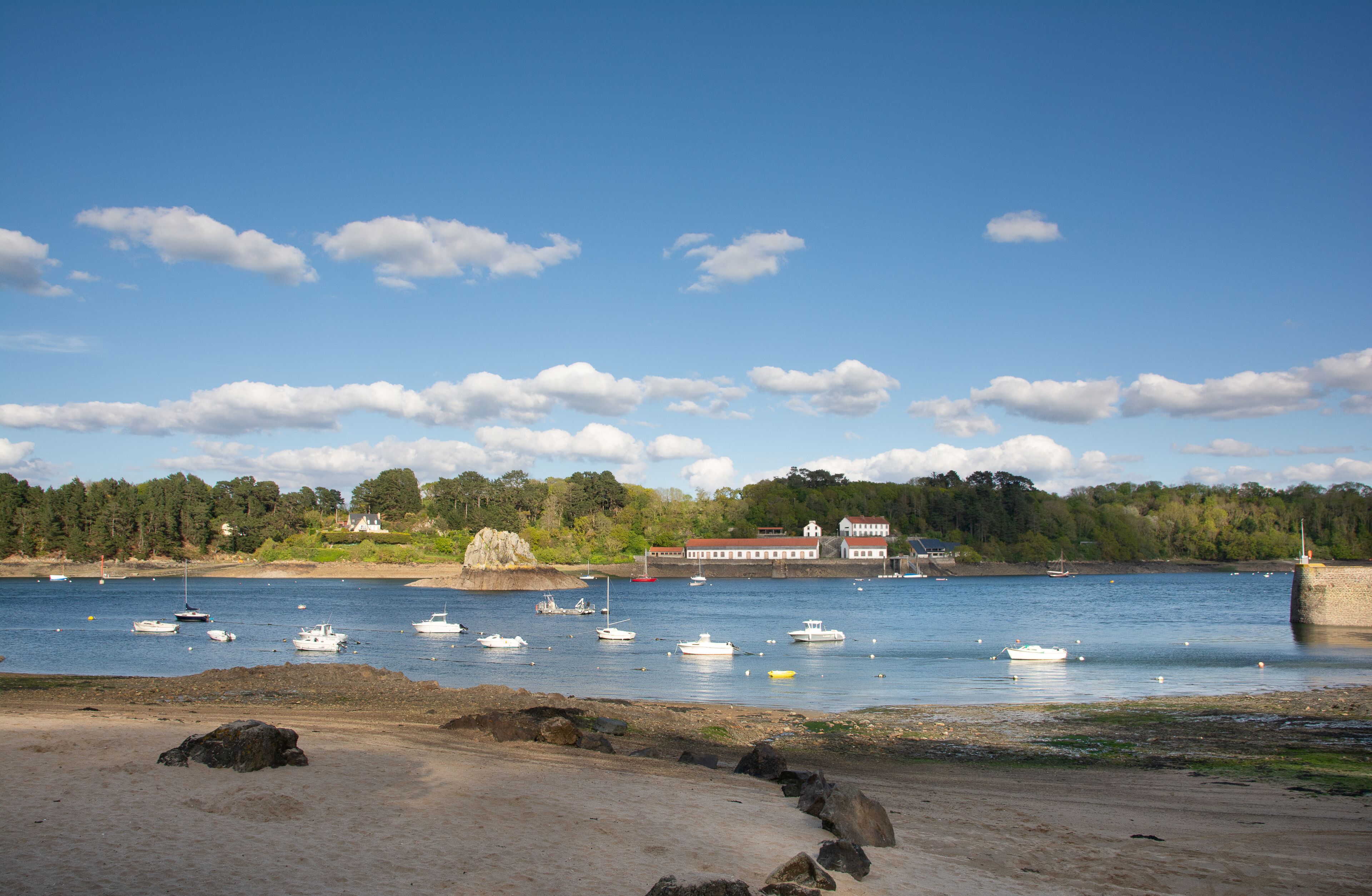 A coastal scene with boats on calm water, a sandy beach, and a building with a red roof in the background.