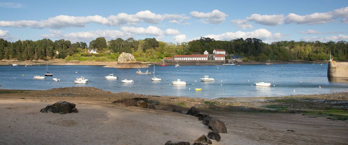A coastal scene with boats on calm water, a sandy beach, and a building with a red roof in the background.