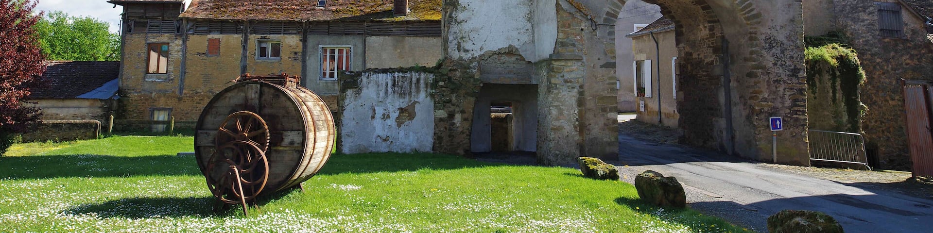 A historic stone building with a large archway and a weathered wooden barrel in a grassy area.
