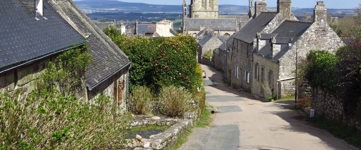 A narrow street lined with stone buildings, a prominent church tower, and a distant view of rolling hills.