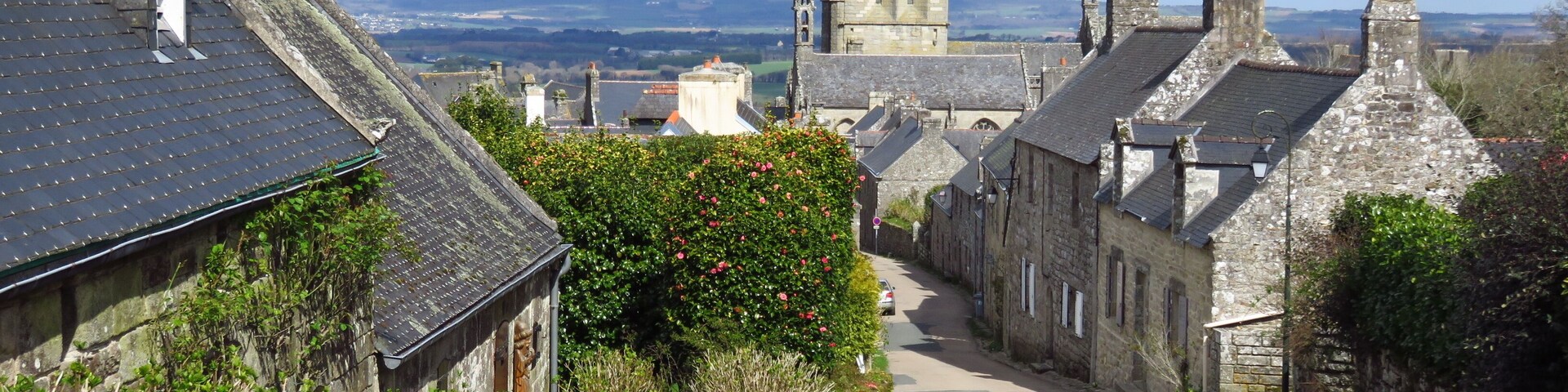 A narrow street lined with stone buildings, a prominent church tower, and a distant view of rolling hills.